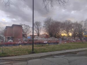 Ground-level view of a commercial demolition project at Dix Park in Raleigh, North Carolina, showing large piles of brick and concrete debris, a partially demolished brick building, perimeter fencing, and heavy demolition equipment as JCE, Inc. completes structural demolition and site clearing.