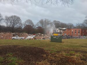 Commercial building demolition in progress with an excavator demolishing a two-story brick structure while water is sprayed for dust control. Demolition debris, perimeter fencing, work vehicles, and heavy equipment are visible as JCE, Inc. completes a safe and controlled demolition project.