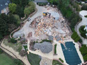 Aerial drone view of a luxury residential demolition project in Charlotte, North Carolina, showing a partially demolished custom home, heavy excavators, demolition debris, a covered swimming pool, and surrounding upscale residences as JCE, Inc. completes safe and controlled structural demolition.